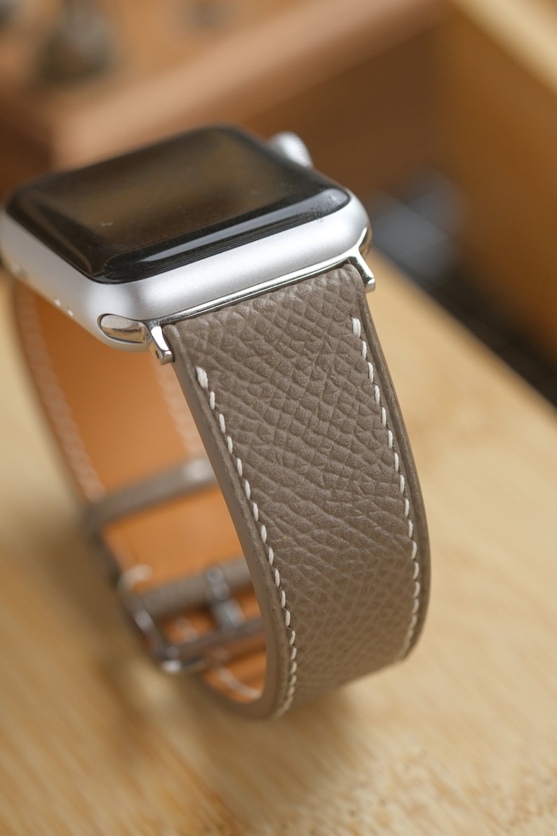 May include: A silver Apple Watch with a taupe leather band. The band has white stitching and a silver buckle. The watch face is black. The leather has a textured appearance.