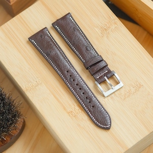 Puede incluir: Dos correas de reloj de cuero marrón con costuras blancas. Las correas tienen un patrón texturizado, similar al del avestruz. Una correa tiene una hebilla plateada.