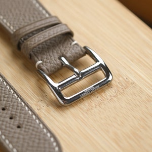 May include: Close-up of a taupe leather watch strap with white stitching and a silver buckle. The buckle is engraved with "BROAD STREET BESPOKE". The strap is on a light brown wooden surface.