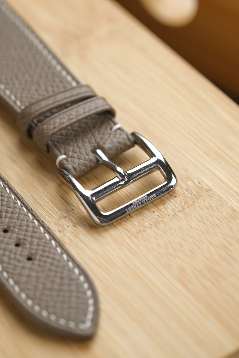 May include: Close-up of a taupe leather watch strap with white stitching and a silver buckle. The buckle is engraved with "BROAD STREET BESPOKE". The strap is on a light brown wooden surface.