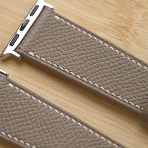 May include: Close-up of a taupe leather watch band with white stitching. The band has a textured surface and a silver-colored metal connector. The watch band is on a light brown wooden surface.