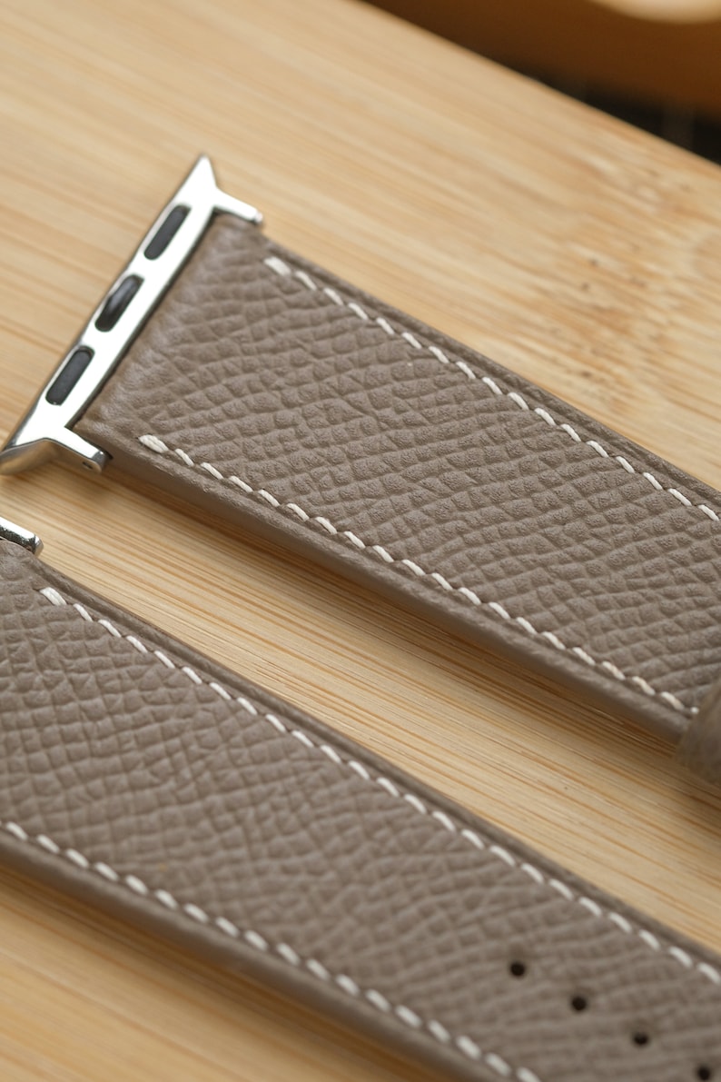 May include: Close-up of a taupe leather watch band with white stitching. The band has a textured surface and a silver-colored metal connector. The watch band is on a light brown wooden surface.