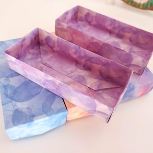 May include: Three rectangular boxes made of paper with a watercolor design. The boxes are stacked on top of each other. The top box is purple with a light pink watercolor design. The middle box is light pink with a light blue watercolor design. The bottom box is light blue with a dark blue watercolor design.