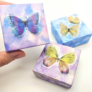May include: Three small square boxes with watercolor backgrounds in shades of blue and purple. Each box features a holographic butterfly sticker. One box is held by a hand. The butterflies have blue, yellow, and purple wings.