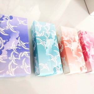 May include: Four small boxes with a gradient design featuring white cranes flying against a background of clouds. The boxes are arranged in a row from left to right, with the colors transitioning from purple to blue, green, orange, and red.
