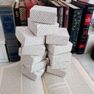 May include: A stack of small, white boxes made from book pages, sitting on top of an open book. The boxes are arranged in a pyramid shape. The book is open to a page with text. There are other books in the background.