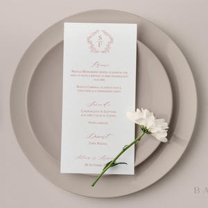 Può includere: Un menu formale con stemma floreale e testo in corsivo, appoggiato su un piatto beige. Un fiore bianco con stelo verde è posto sul piatto. Il menu presenta le iniziali "S" e "F".