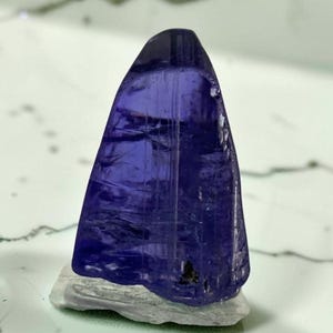 May include: A translucent, deep violet-blue tanzanite crystal with a triangular shape and visible internal striations, resting on a light grey, textured mineral matrix base. The crystal displays varying shades of blue and purple throughout.