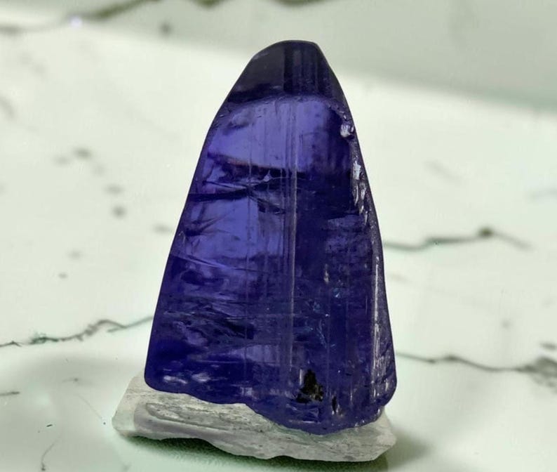 May include: A translucent, deep violet-blue tanzanite crystal with a triangular shape and visible internal striations, resting on a light grey, textured mineral matrix base. The crystal displays varying shades of blue and purple throughout.