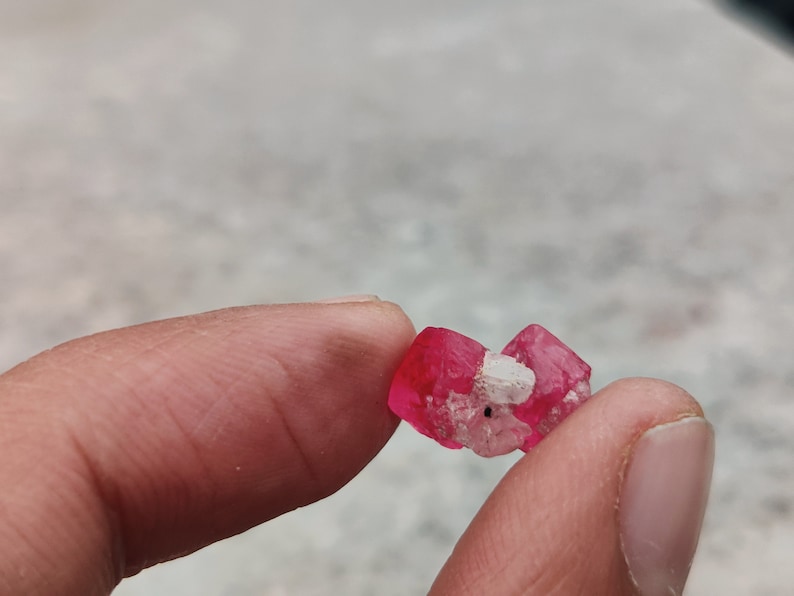 May include: A small, pink, crystalline mineral with a white, cloudy center. The mineral is being held in a hand.