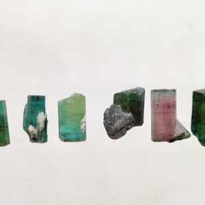 May include: A collection of six green, pink, and black tourmaline crystals. The crystals are all different shapes and sizes, and they are arranged in a row on a white background.