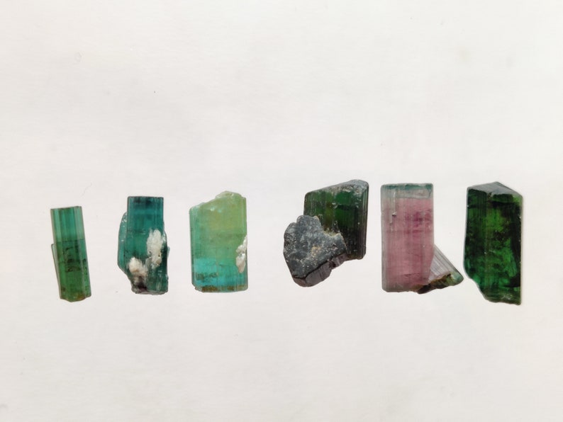 May include: A collection of six green, pink, and black tourmaline crystals. The crystals are all different shapes and sizes, and they are arranged in a row on a white background.