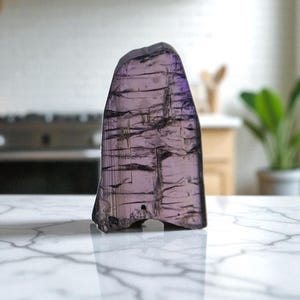 May include: A translucent, irregularly shaped, purple gemstone with dark linear inclusions. The polished surface reflects light, creating a shimmering effect. The stone is displayed on a white marble countertop, showcasing its natural beauty.