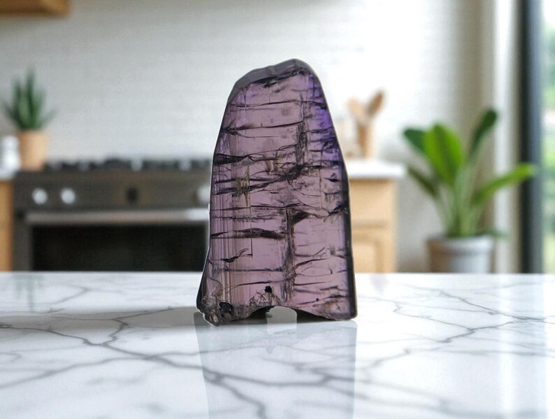 May include: A translucent, irregularly shaped, purple gemstone with dark linear inclusions. The polished surface reflects light, creating a shimmering effect. The stone is displayed on a white marble countertop, showcasing its natural beauty.