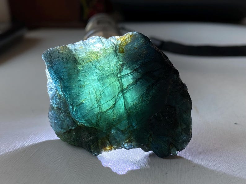 K&ouml;nnte beinhalten: Ein nat&uuml;rlich geformter Edelstein mit einer lebendigen Farbmischung. Der Stein zeigt tiefes T&uuml;rkis, Blau und einen Hauch von Gelbgr&uuml;n, mit sichtbaren inneren Strukturen. Die Oberfl&auml;che ist strukturiert und uneben, wodurch ein schimmernder Effekt entsteht, der das Licht einf&auml;ngt.