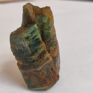 May include: A green and brown crystal with a rough, textured surface. The crystal is shaped like a tall, thin tower.
