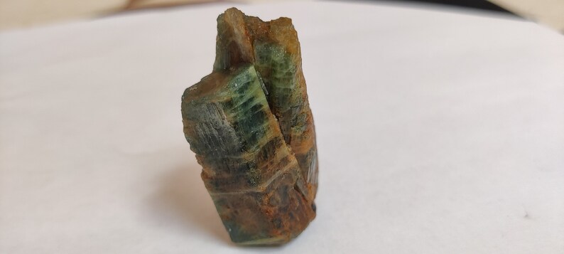 May include: A green and brown crystal with a rough, textured surface. The crystal is shaped like a tall, thin tower.