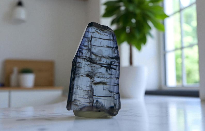 May include: A tall, irregularly shaped, translucent blue crystal with dark linear inclusions. The crystal is set against a blurred background with a white pot, a plant, and a window. The crystal's base is a pale yellow color.