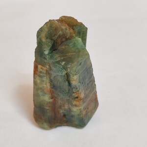 May include: A green and brown crystal with a rough, natural texture. The crystal is shaped like a tower and has multiple facets.