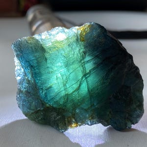 K&ouml;nnte beinhalten: Ein nat&uuml;rlich geformter Edelstein mit einer auff&auml;lligen Farbgebung. Der Stein zeigt eine Mischung aus tiefem Teal, T&uuml;rkis und goldenen Akzenten. Die Oberfl&auml;che hat ein strukturiertes, kristallines Aussehen mit sichtbaren Linien und Mustern.