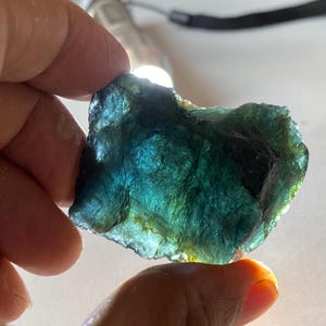 K&ouml;nnte beinhalten: Ein grob geschnittener, durchscheinender Edelstein mit einer Mischung aus Blau- und Gr&uuml;nt&ouml;nen. Der Stein wird vor eine Lichtquelle gehalten, wodurch seine innere Struktur und Farbvariationen sichtbar werden. Die Kanten sind uneben, was auf einen nat&uuml;rlichen, unpolierten Zustand hindeutet.