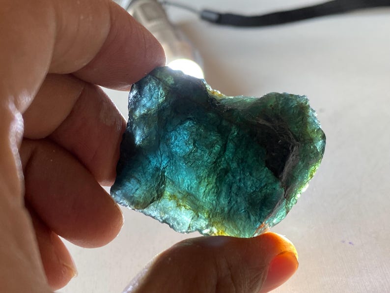 K&ouml;nnte beinhalten: Ein grob geschnittener, durchscheinender Edelstein mit einer Mischung aus Blau- und Gr&uuml;nt&ouml;nen. Der Stein wird vor eine Lichtquelle gehalten, wodurch seine innere Struktur und Farbvariationen sichtbar werden. Die Kanten sind uneben, was auf einen nat&uuml;rlichen, unpolierten Zustand hindeutet.