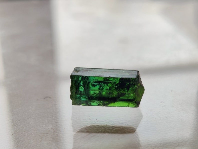 May include: A dark green, faceted, rectangular gemstone with a slight curve on one side.