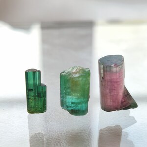 May include: Three tourmaline crystals, one green, one green and blue, and one pink and green. The crystals are on a white surface.