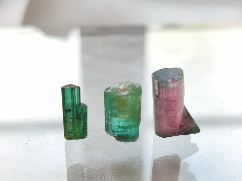 May include: Three tourmaline crystals, one green, one green and blue, and one pink and green. The crystals are on a white surface.