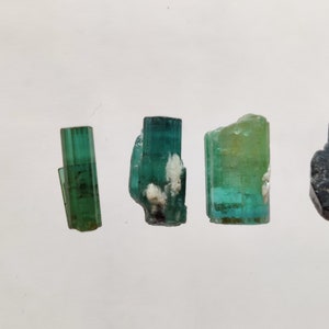 May include: Four green and blue gemstone crystals on a white background. The crystals are all different sizes and shapes.