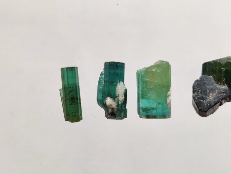 May include: Four green and blue gemstone crystals on a white background. The crystals are all different sizes and shapes.