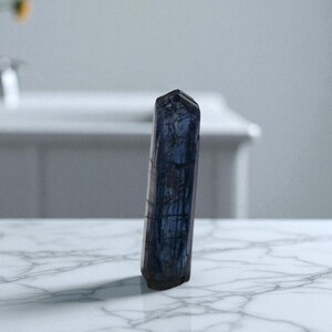 May include: A dark blue crystal stands upright on a white marble surface. The crystal has a faceted top and a textured surface. The background is blurred, showing a white cabinet and a vase with flowers.