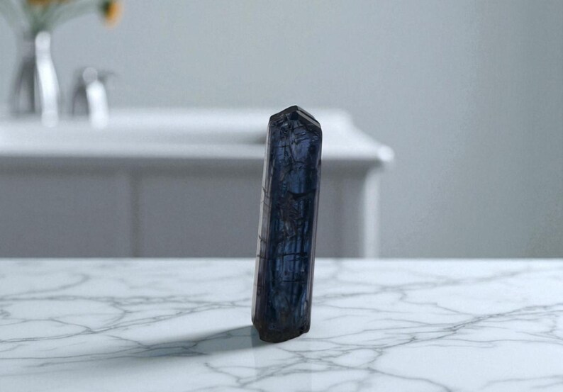 May include: A dark blue crystal stands upright on a white marble surface. The crystal has a faceted top and a textured surface. The background is blurred, showing a white cabinet and a vase with flowers.