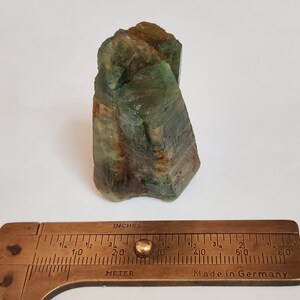 May include: A green and brown crystal cluster with a rough, natural texture. The crystal is approximately 2 inches tall and is measured with a brass ruler.