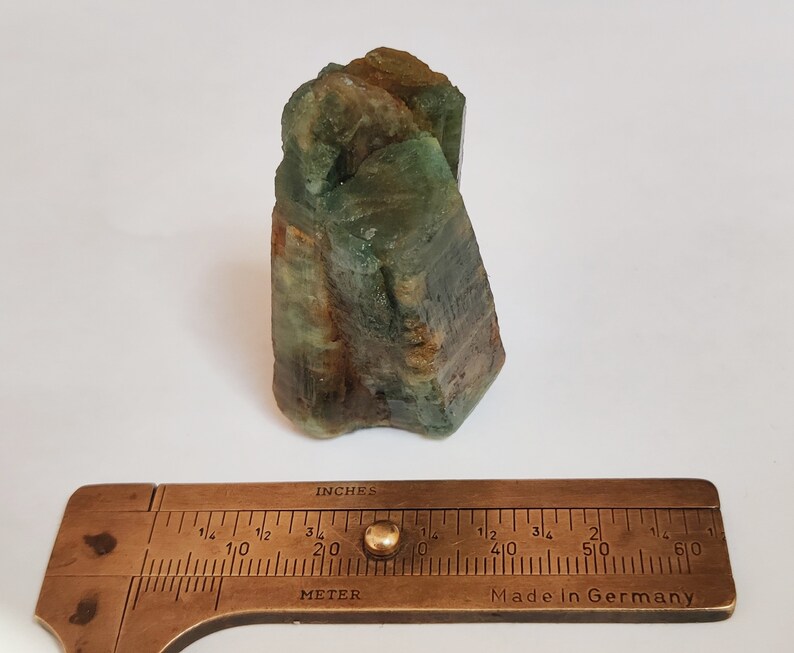 May include: A green and brown crystal cluster with a rough, natural texture. The crystal is approximately 2 inches tall and is measured with a brass ruler.