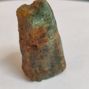 May include: A rough, green and brown gemstone with a pointed tip. The gemstone has a natural, unpolished look.