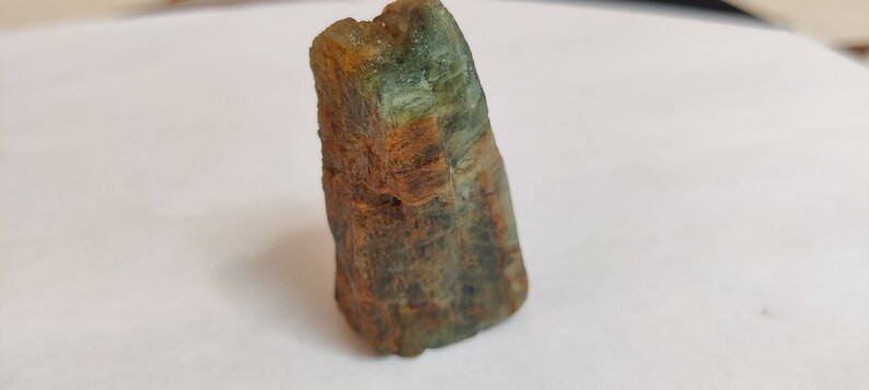 May include: A rough, green and brown gemstone with a pointed tip. The gemstone has a natural, unpolished look.