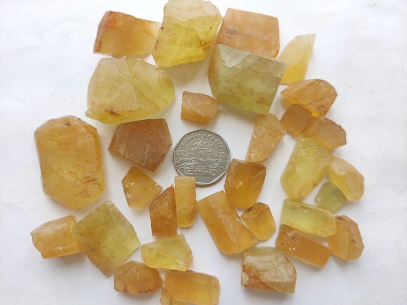 Puede incluir: Una colecci&oacute;n de cristales amarillos y naranjas, algunos con un tono verdoso, dispuestos sobre una superficie blanca. Una moneda de 25 centavos de d&oacute;lar estadounidense se incluye para la escala.
