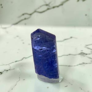 May include: A single, vertically oriented tanzanite crystal featuring a deep violet-blue hue. The gemstone displays a translucent, faceted structure with visible internal inclusions and a polished, flat base that allows it to stand upright.