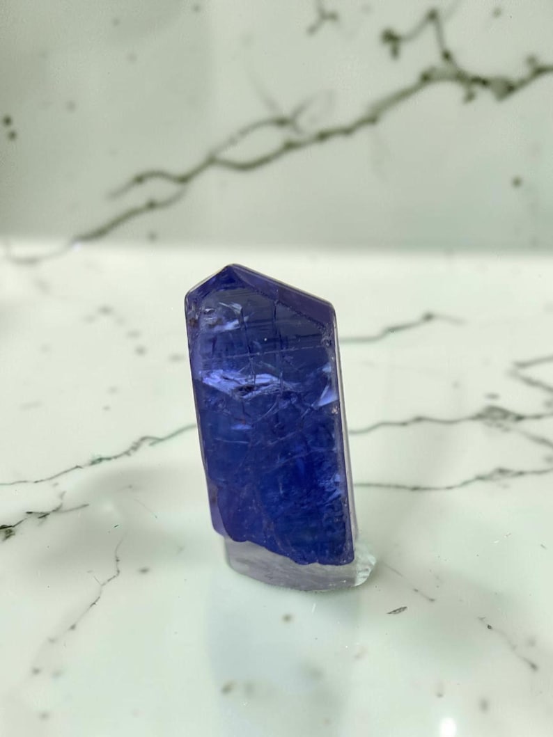 May include: A single, vertically oriented tanzanite crystal featuring a deep violet-blue hue. The gemstone displays a translucent, faceted structure with visible internal inclusions and a polished, flat base that allows it to stand upright.