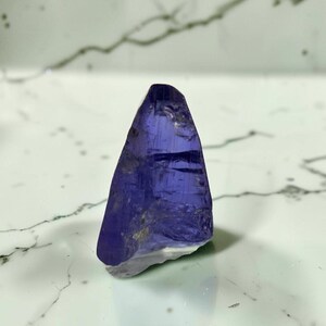 May include: A raw, translucent tanzanite crystal with a deep violet-blue hue. The stone features a triangular, pointed shape with natural facets and internal inclusions, displaying a polished, glassy texture on its primary surfaces.