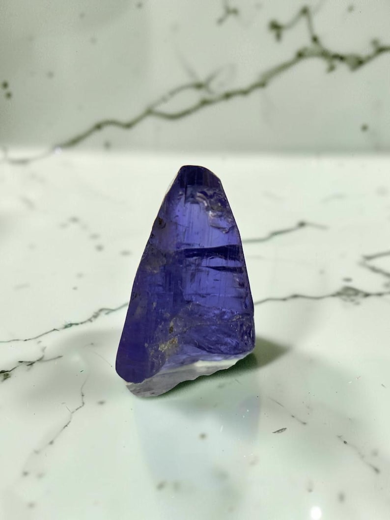May include: A raw, translucent tanzanite crystal with a deep violet-blue hue. The stone features a triangular, pointed shape with natural facets and internal inclusions, displaying a polished, glassy texture on its primary surfaces.