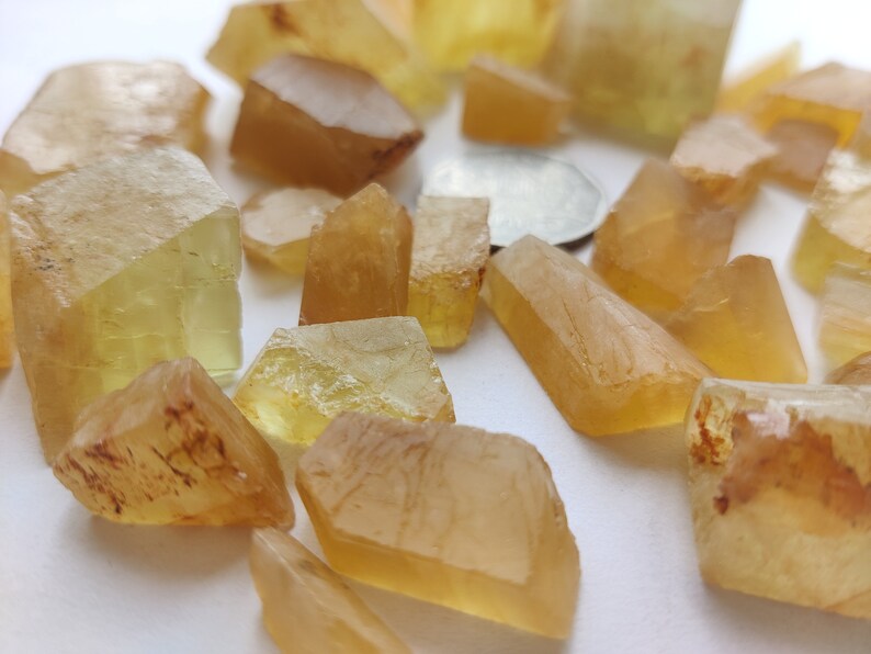 Puede incluir: Una colecci&oacute;n de cristales de color amarillo y &aacute;mbar, algunos con un ligero tono rojizo, esparcidos sobre una superficie blanca.