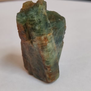 May include: A green and brown rough, natural, crystal specimen. The crystal has a striated, layered appearance.