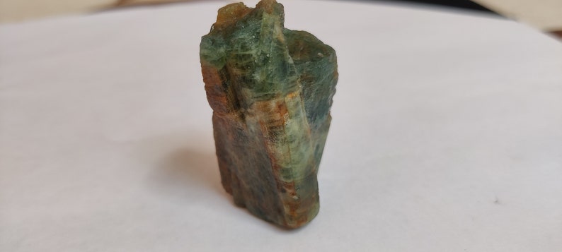 May include: A green and brown rough, natural, crystal specimen. The crystal has a striated, layered appearance.
