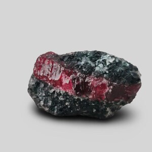 May include: A raw mineral specimen featuring a vibrant, translucent red crystal band embedded within a dark, textured, charcoal-colored matrix. The red section displays a faceted, crystalline structure with visible internal depth and light reflection.