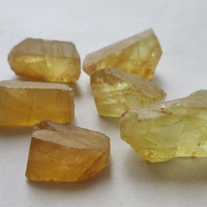 Puede incluir: Seis piedras preciosas talladas en bruto, facetadas, de color amarillo. Las piedras preciosas son transl&uacute;cidas y tienen un ligero tono verde. Las piedras preciosas est&aacute;n dispuestas en un racimo sobre una superficie blanca.