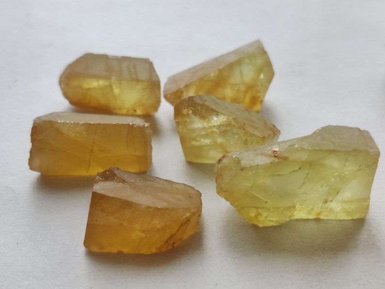 Puede incluir: Seis piedras preciosas talladas en bruto, facetadas, de color amarillo. Las piedras preciosas son transl&uacute;cidas y tienen un ligero tono verde. Las piedras preciosas est&aacute;n dispuestas en un racimo sobre una superficie blanca.