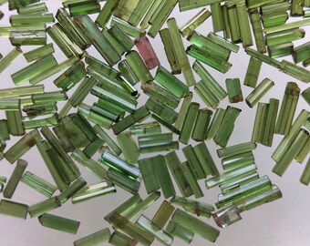 May include: A collection of green tourmaline crystals, also known as green gemstones, in various sizes and shapes. The crystals are arranged on a white surface.