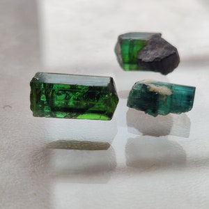 May include: Three green gemstones, two of which are long and rectangular, and one is shorter and more rounded. The gemstones are on a white surface.